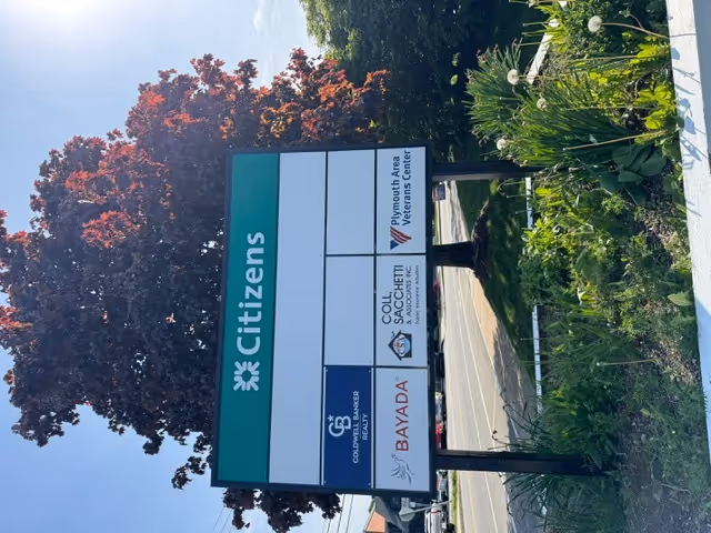 Outdoor signboard with BAYADA logo and other tenants beside trees near a sidewalk.