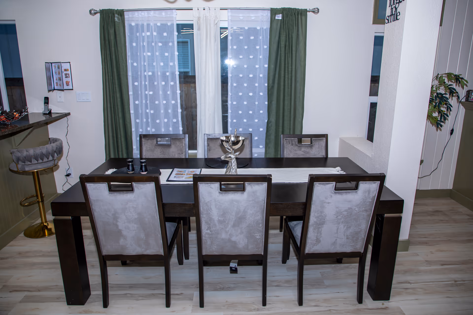 Dark dining table with six upholstered chairs in a bright room with green curtains.