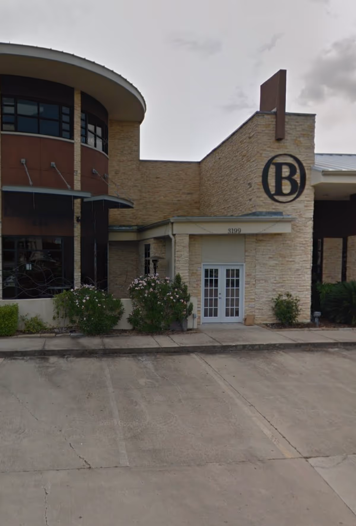 Front exterior of beige brick senior facility with a circular black B logo and glass doors.