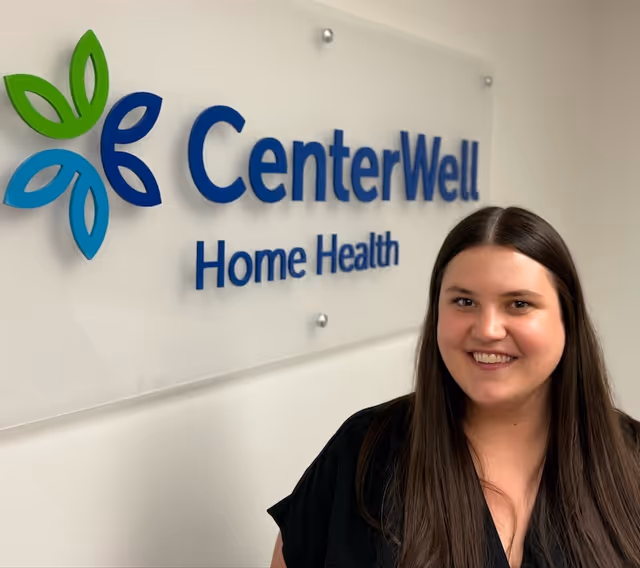Smiling woman stands beside a CenterWell Home Health sign on an interior wall.