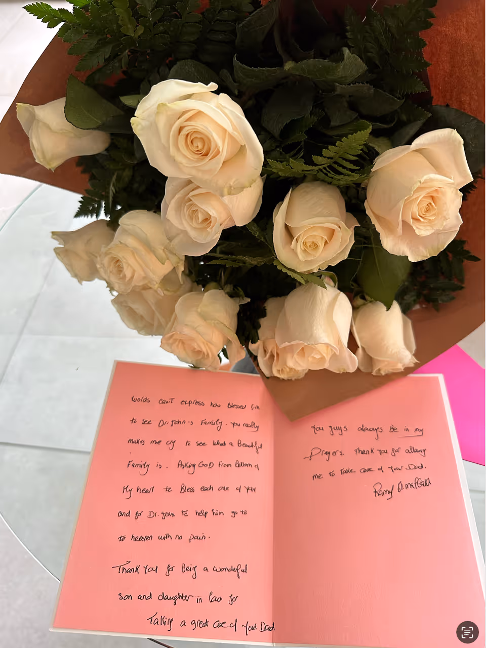 Bouquet of ivory roses beside a pink card on a glass-topped round table.