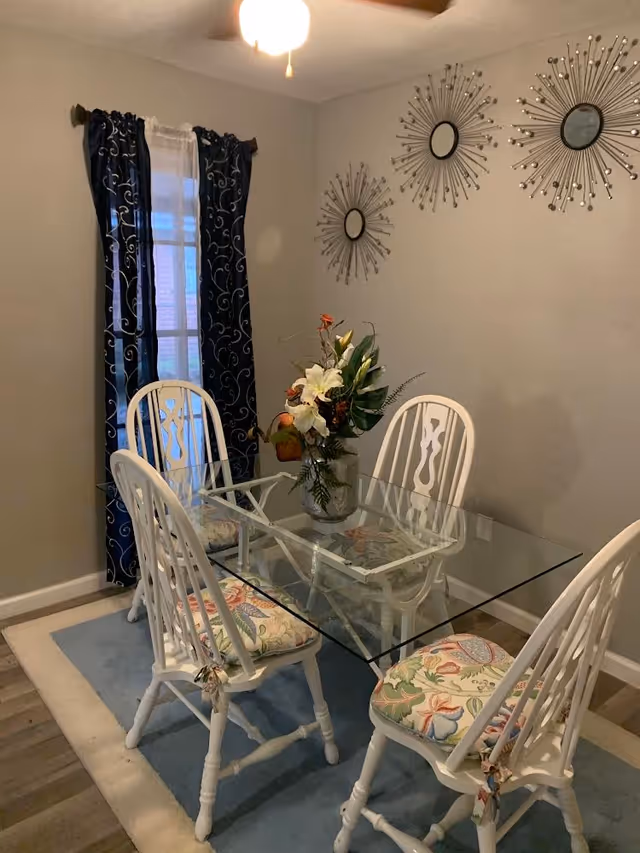 Bright dining room with glass table, white chairs, floral cushions, and sunburst wall decor.