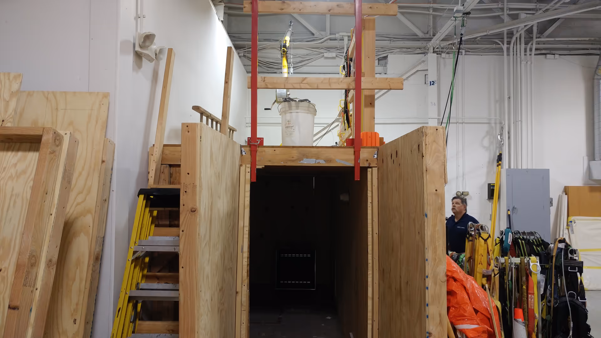 Interior workshop with plywood, ladders, scaffolding, and a man beside construction gear near a dark doorway.