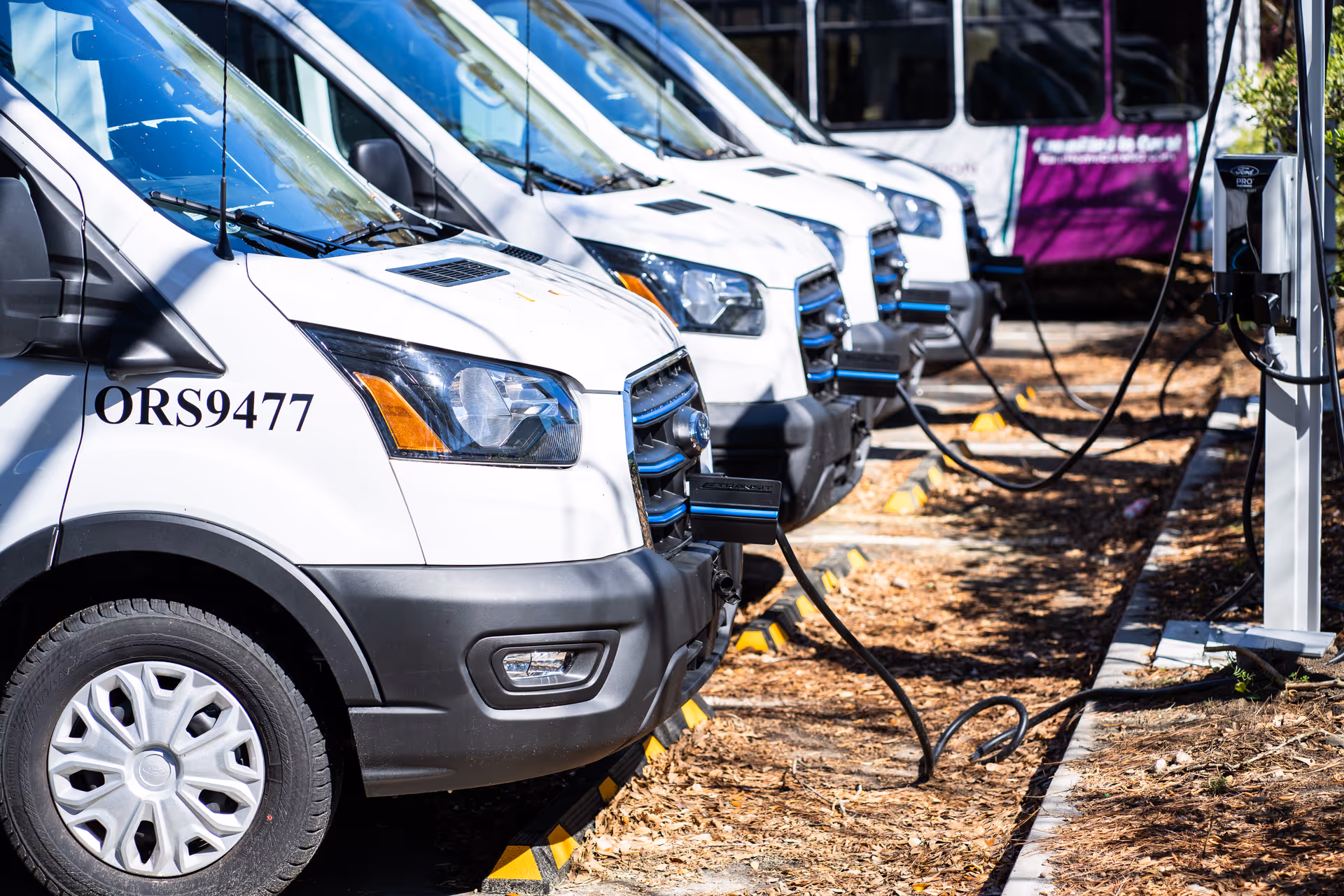 Row of white shuttle vans parked outside, connected to charging stations in a lot.