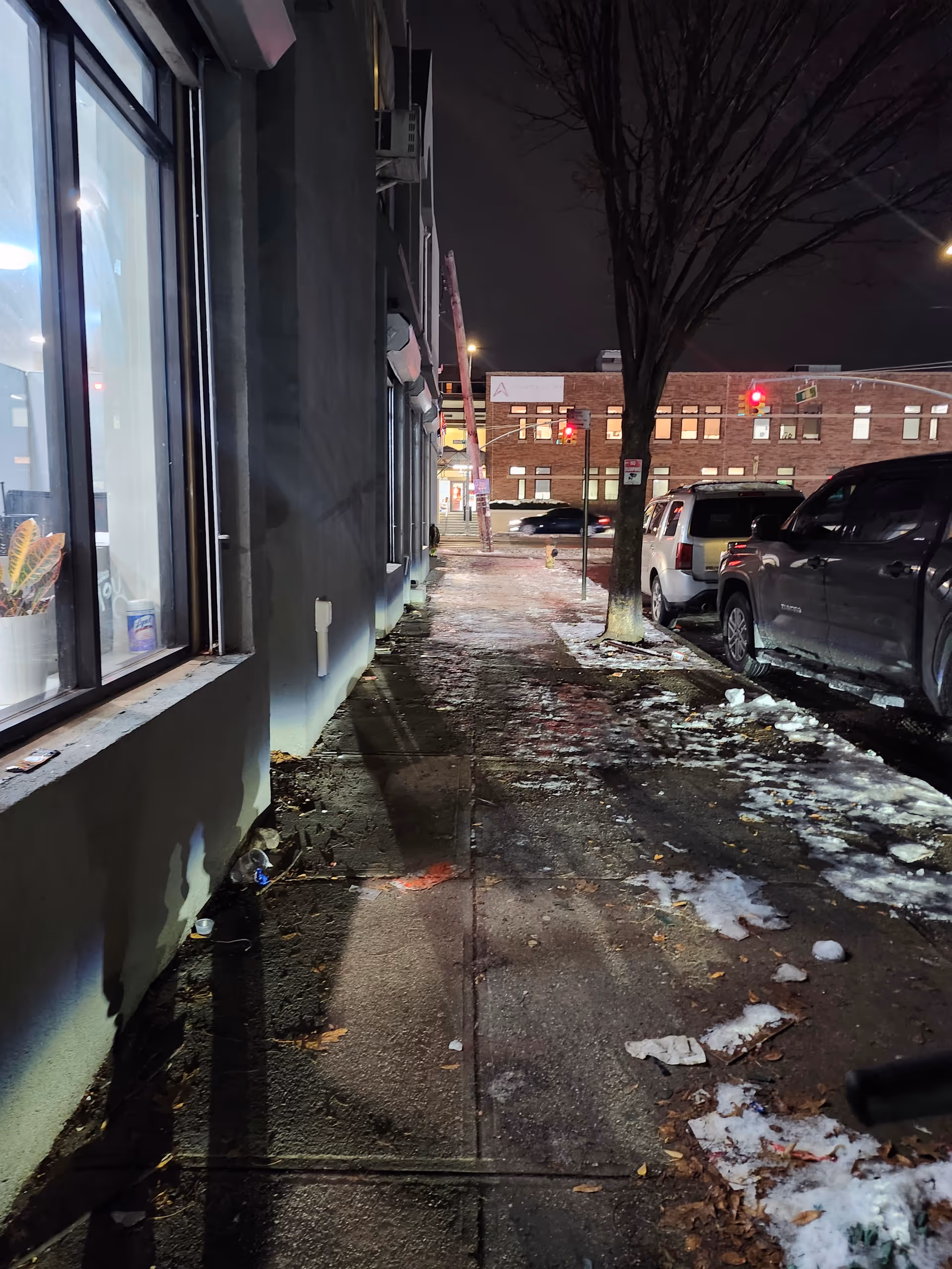 Nighttime sidewalk beside a building with snow, parked cars, and bare trees.