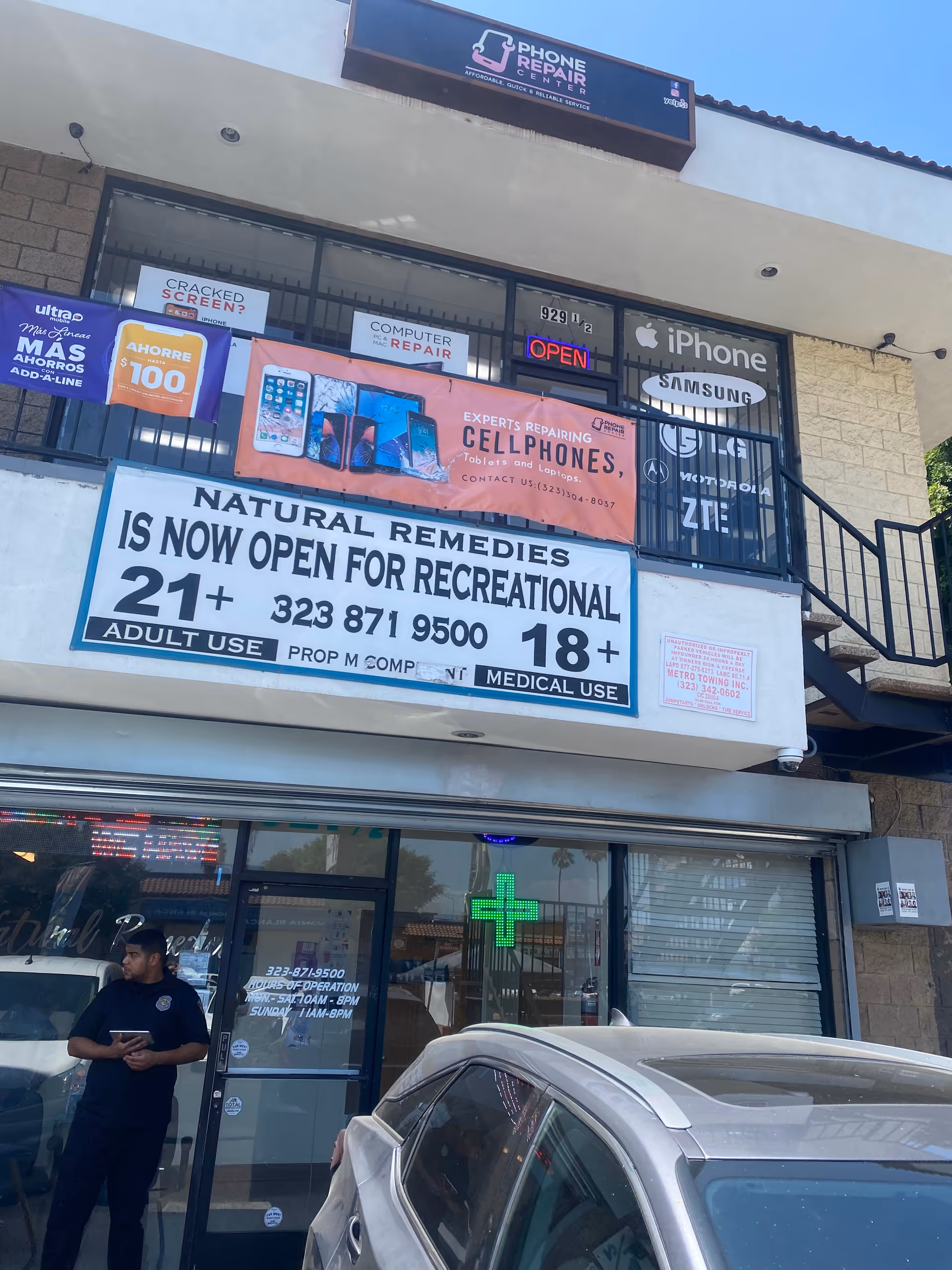 Two-story storefronts with banners advertising Natural Remedies and a green pharmacy cross beside parked cars.