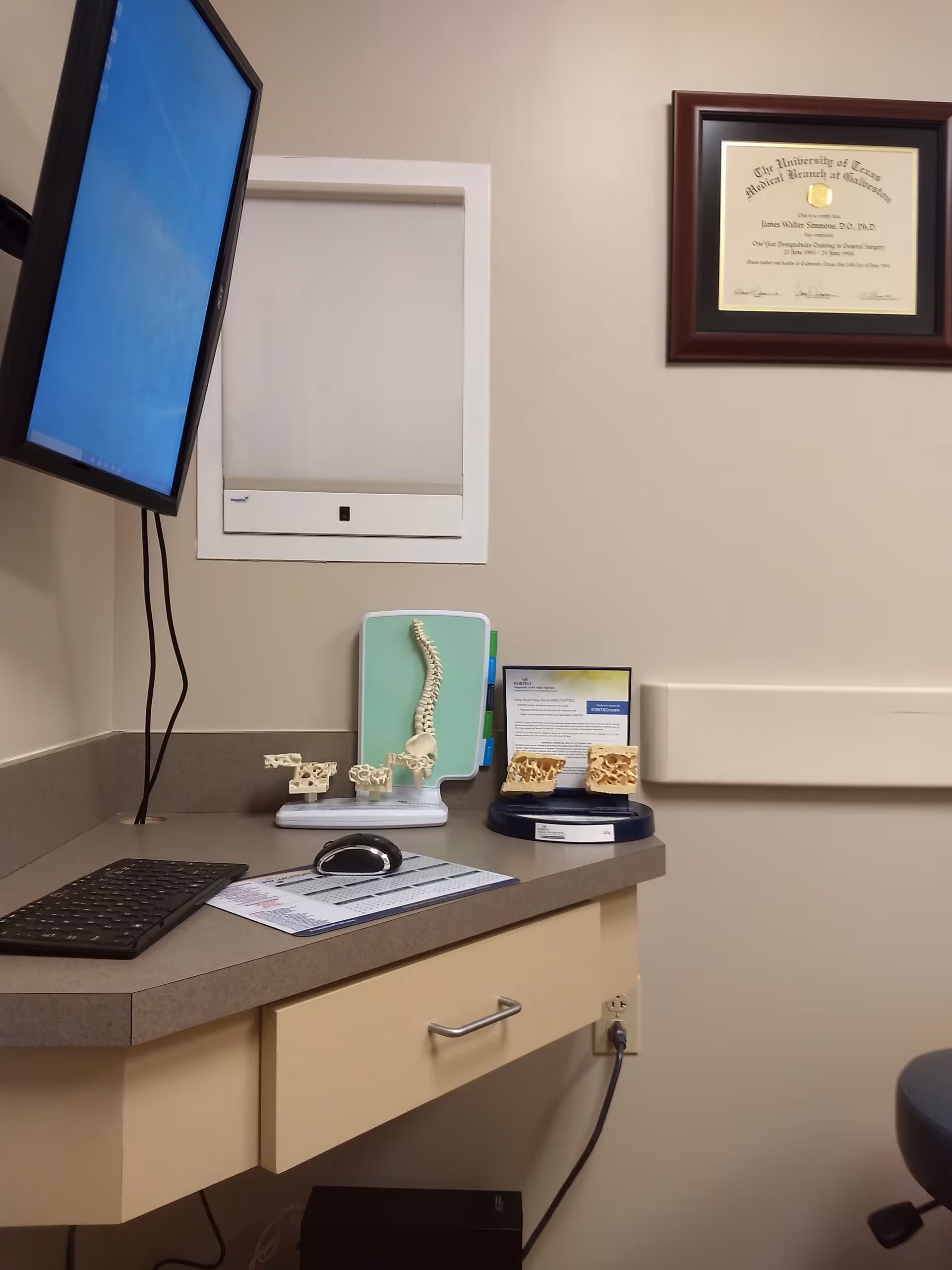 Medical exam room with a desk, monitor, spine models, and a diploma on the wall.