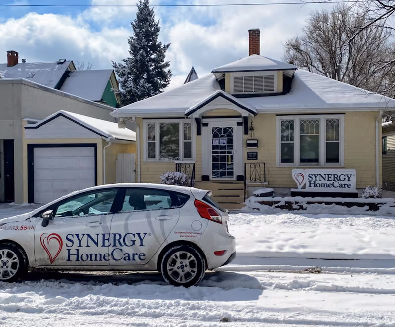 Snow-covered single-story house with SYNERGY HomeCare sign and branded car parked out front