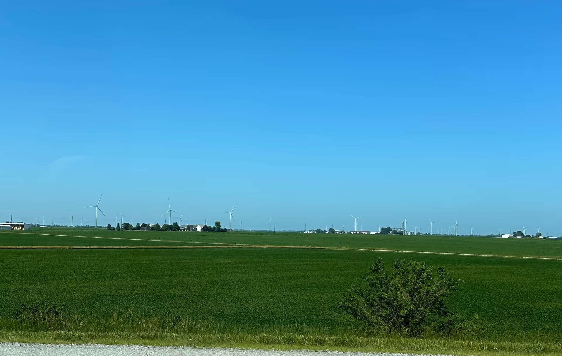 Wide rural landscape with green fields and wind turbines beneath a clear blue sky.