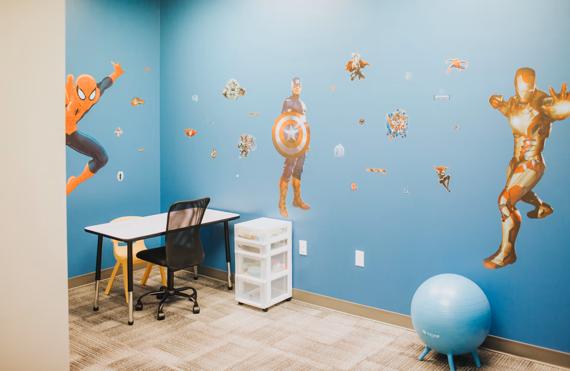 Interior playroom with blue walls, superhero decals, a desk, chair, and storage unit.