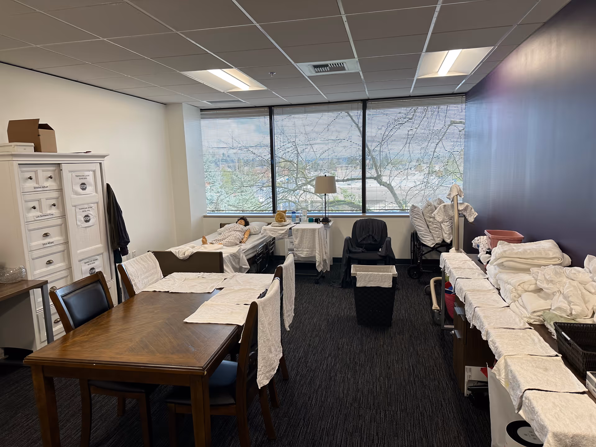 Room with several beds along a windowed wall, a dining table, and medical supplies.