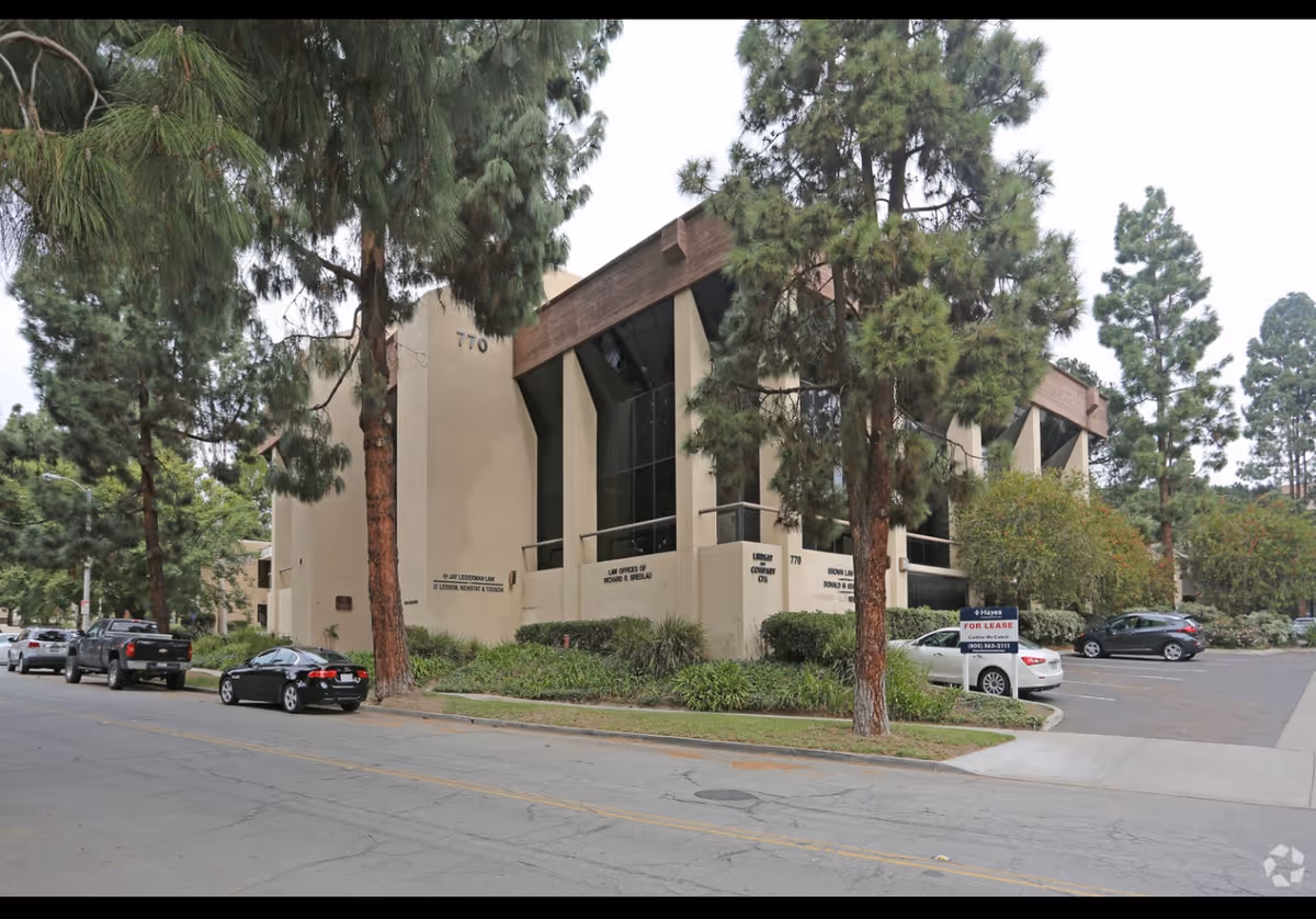 Exterior view of a beige building with tall trees and a parking lot.