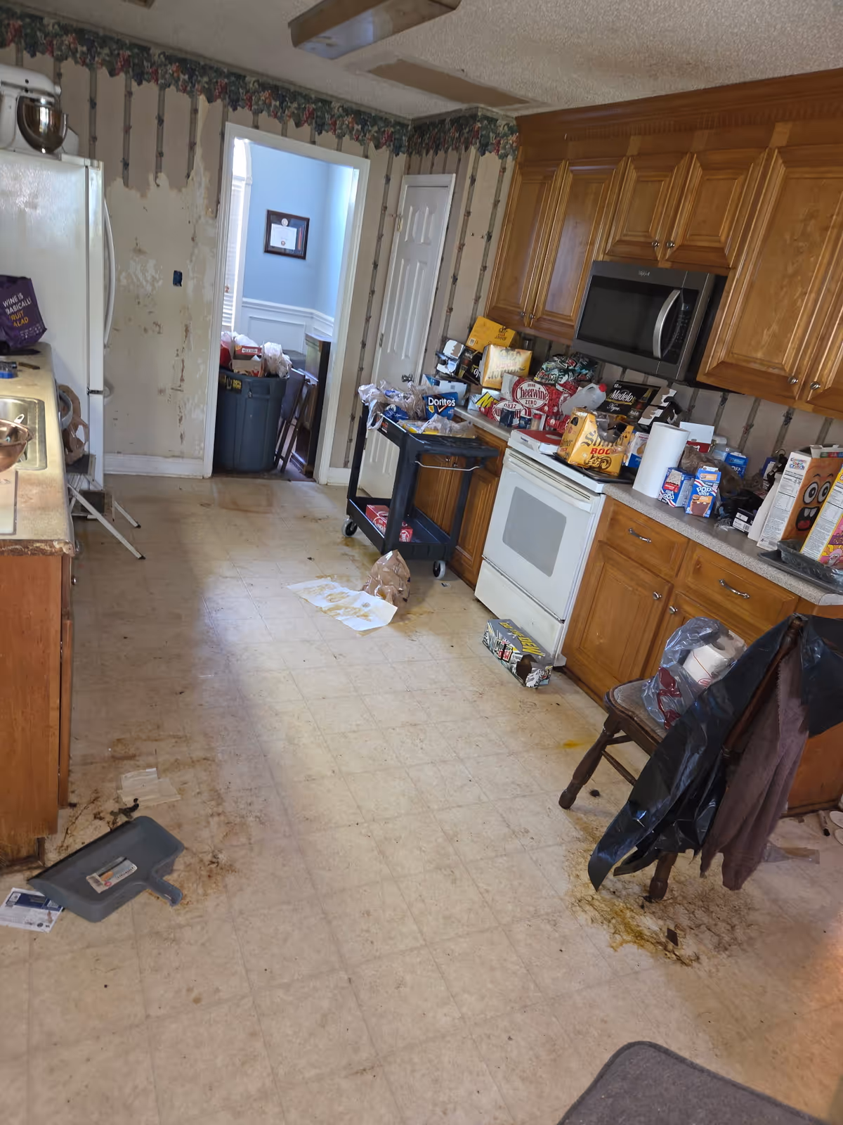 Cluttered residential kitchen with wooden cabinets, appliances, and trash on the floor.