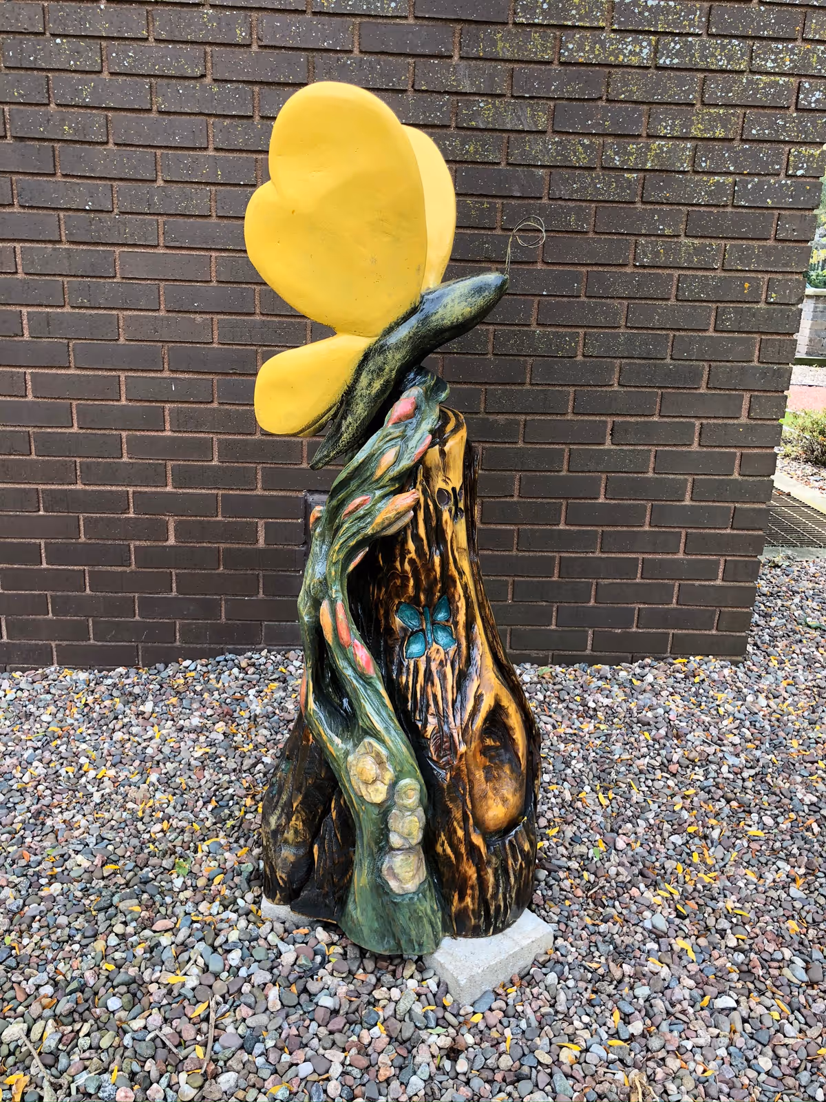 Colorful outdoor sculpture of a tree trunk with a yellow butterfly beside brick wall.