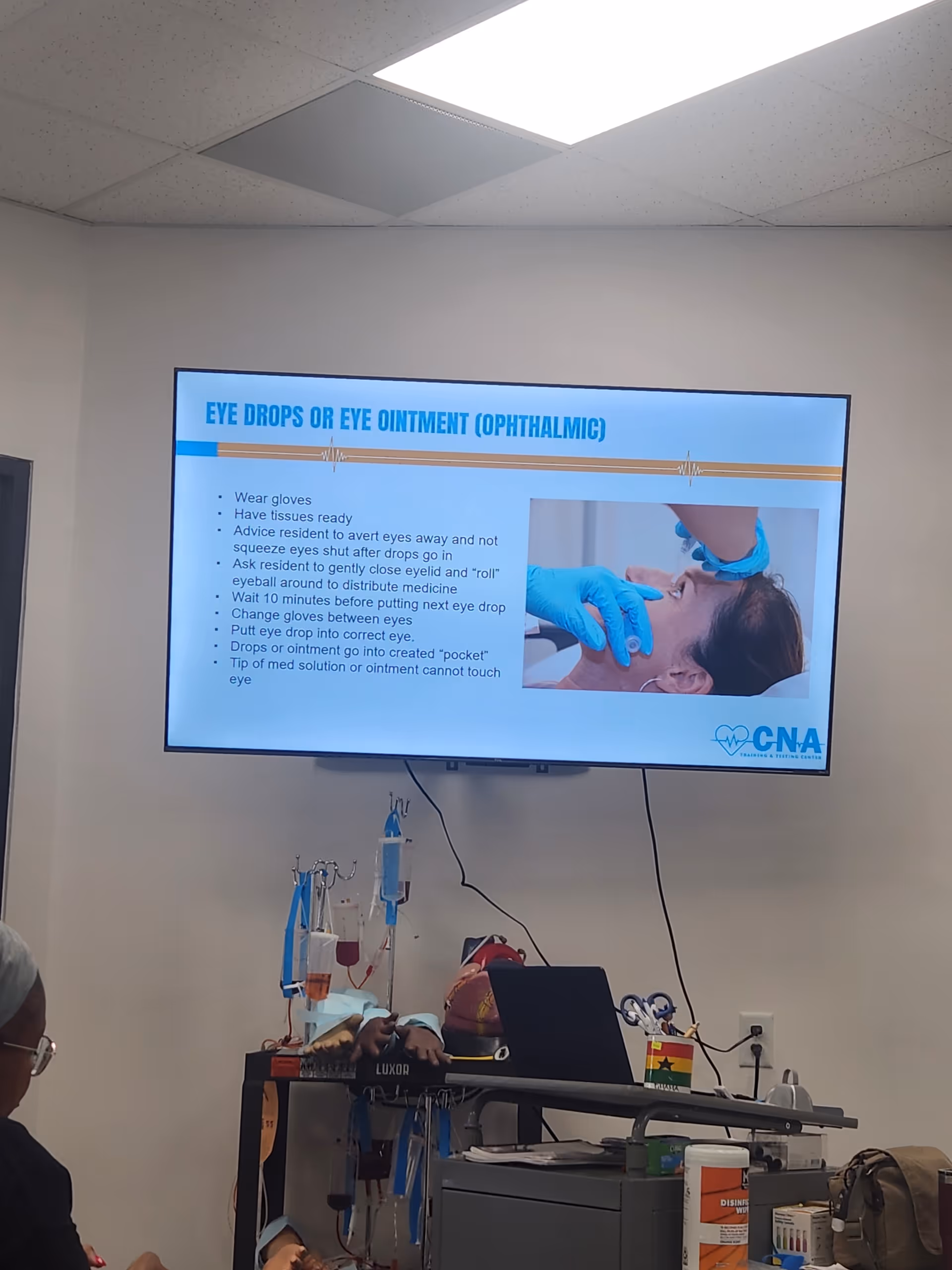 Wall-mounted screen displays ophthalmic instructions beside medical equipment in a training room.