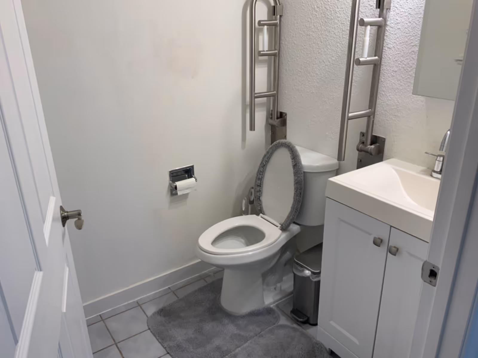 Accessible bathroom with toilet, wall-mounted grab bars, sink, cabinet, and gray rug.