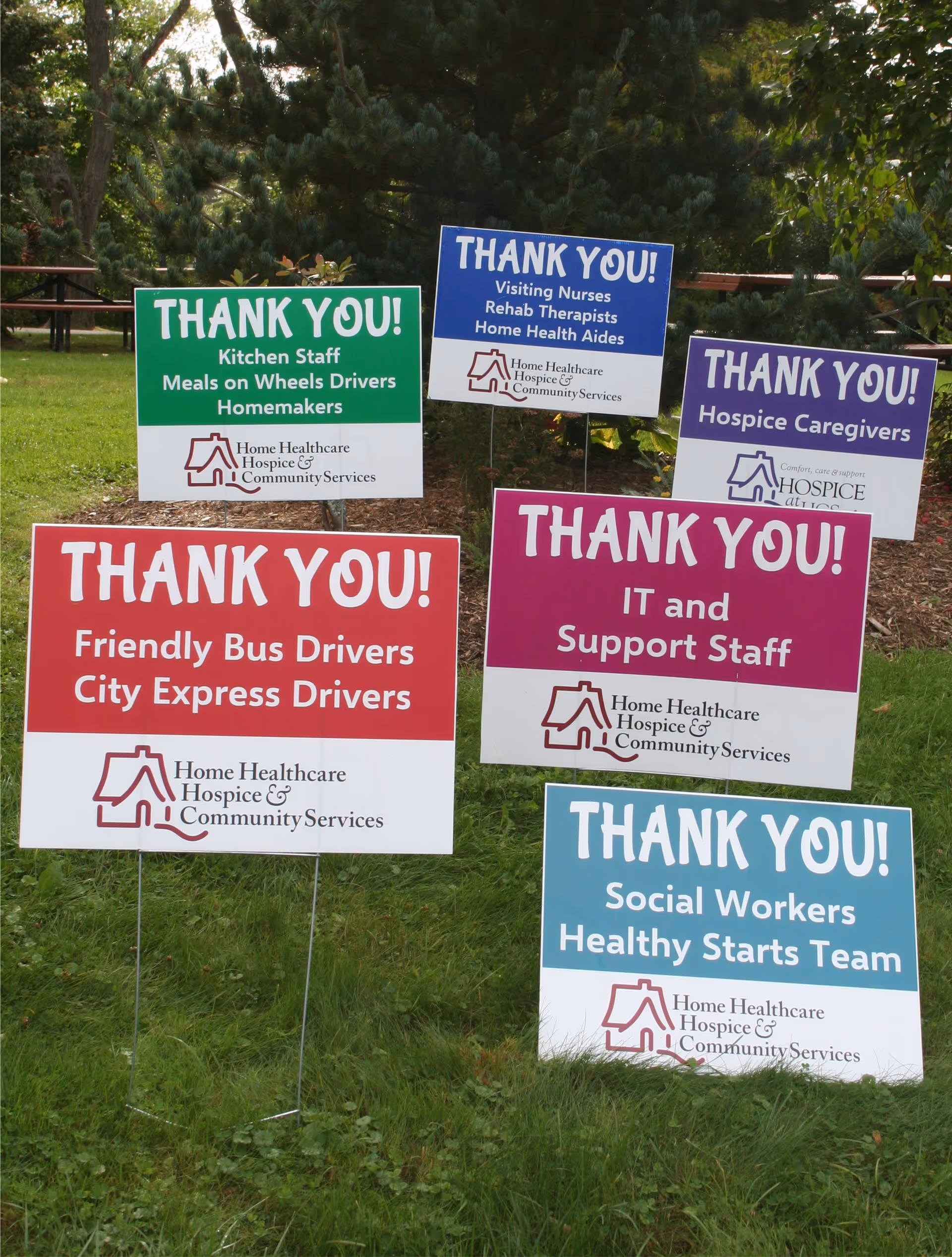 Outdoor scene with multiple colorful thank-you signs on a grassy area near trees.