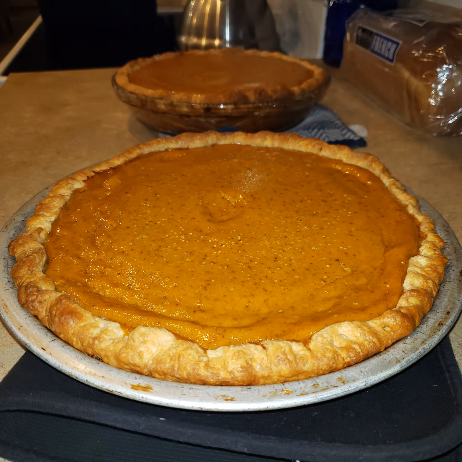 Two pumpkin pies cooling on a kitchen counter with bread in the background
