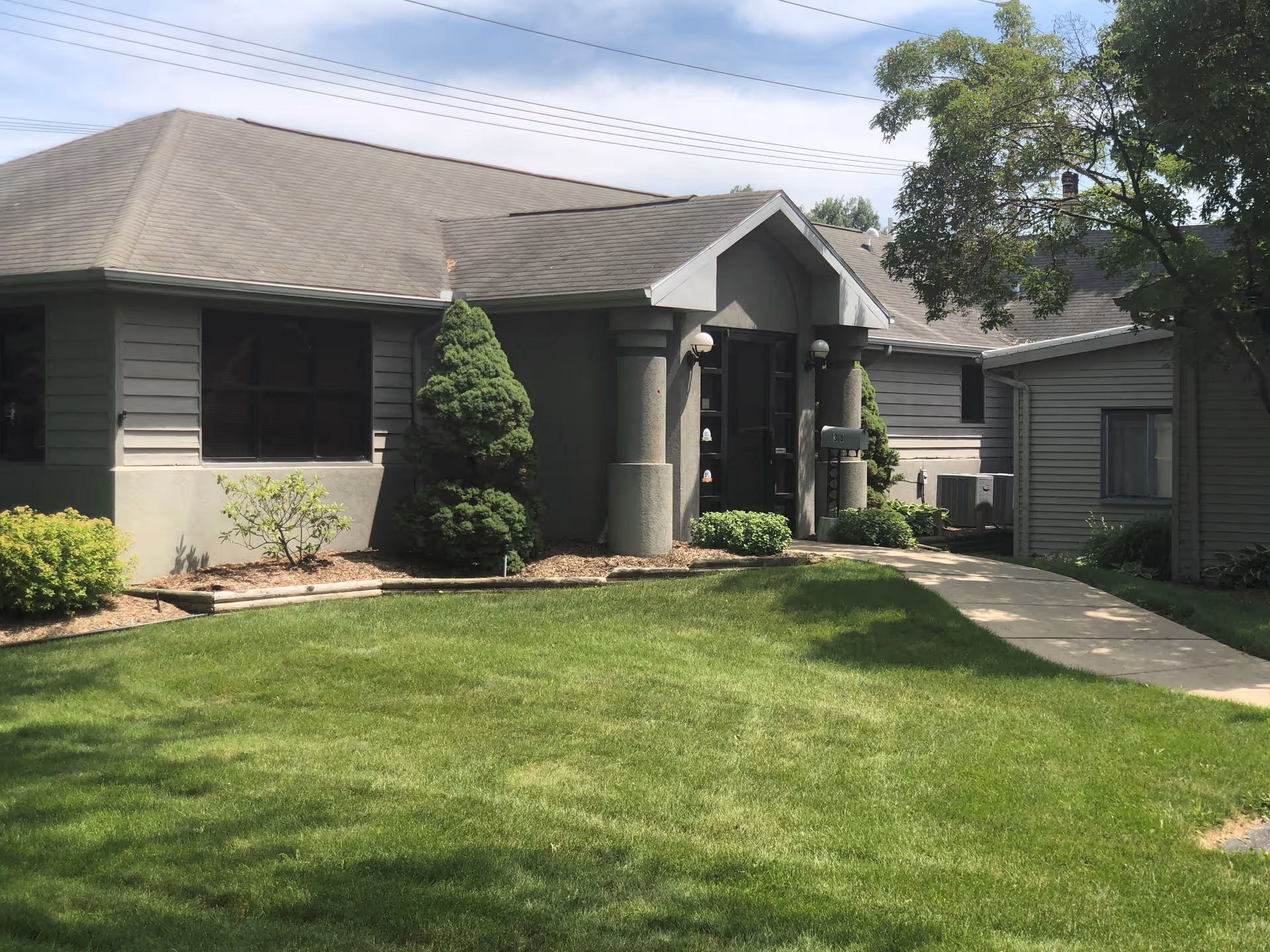 Front view of a single-story gray building with a peaked entrance, columns, and a green lawn.
