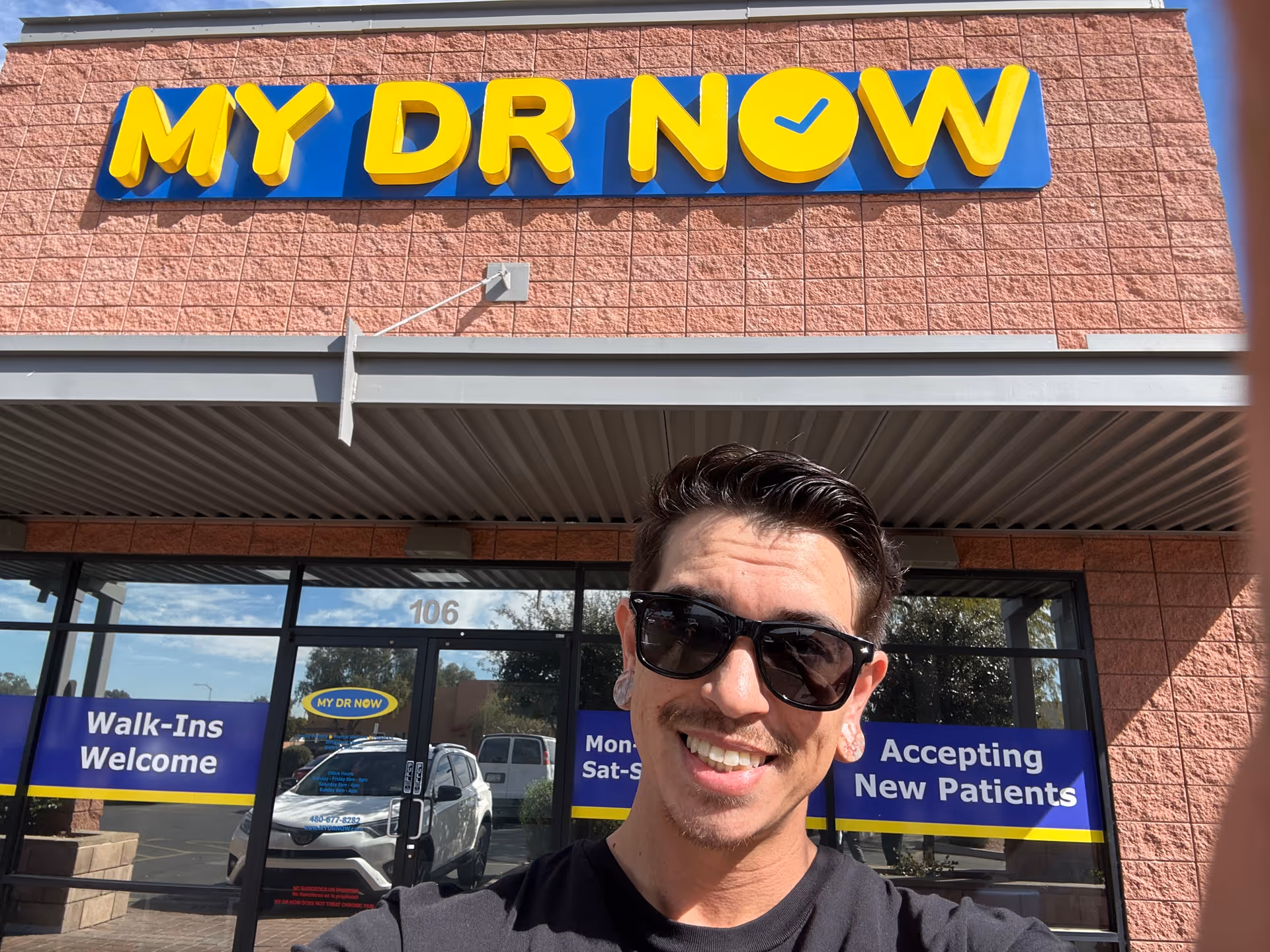 Smiling man wearing sunglasses takes a selfie in front of a MY DR NOW clinic storefront.