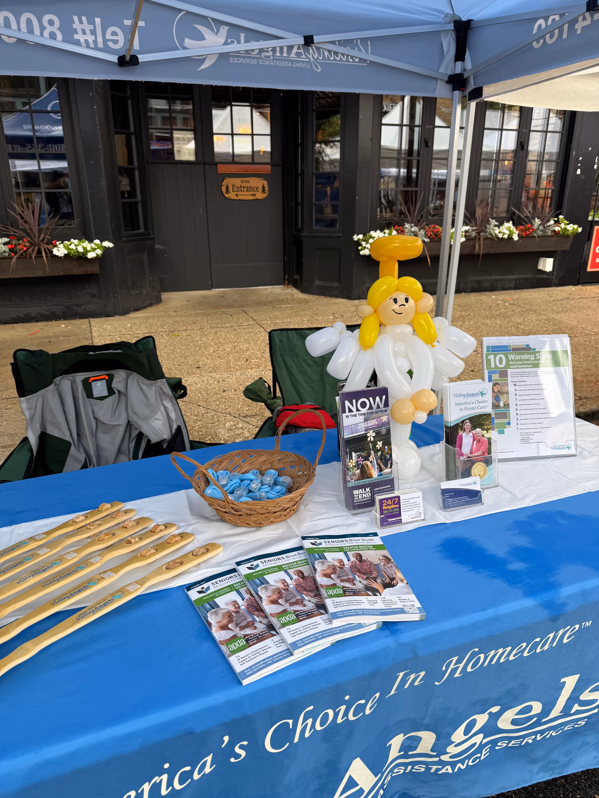 Outdoor promotional table under a canopy with brochures and a balloon figure near an entrance.