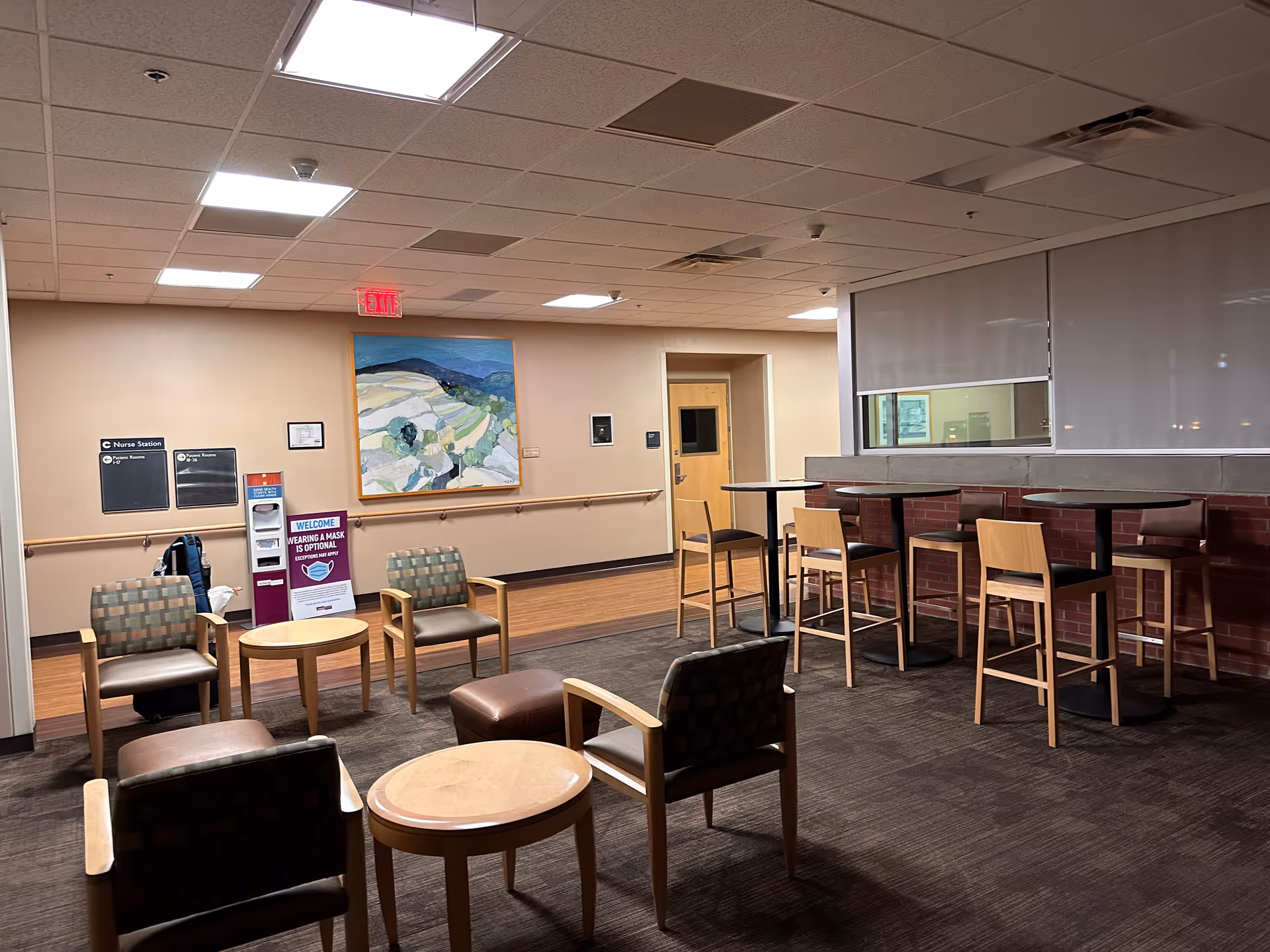 Empty hospital lounge with chairs, round tables, and a high-top counter near a service window.