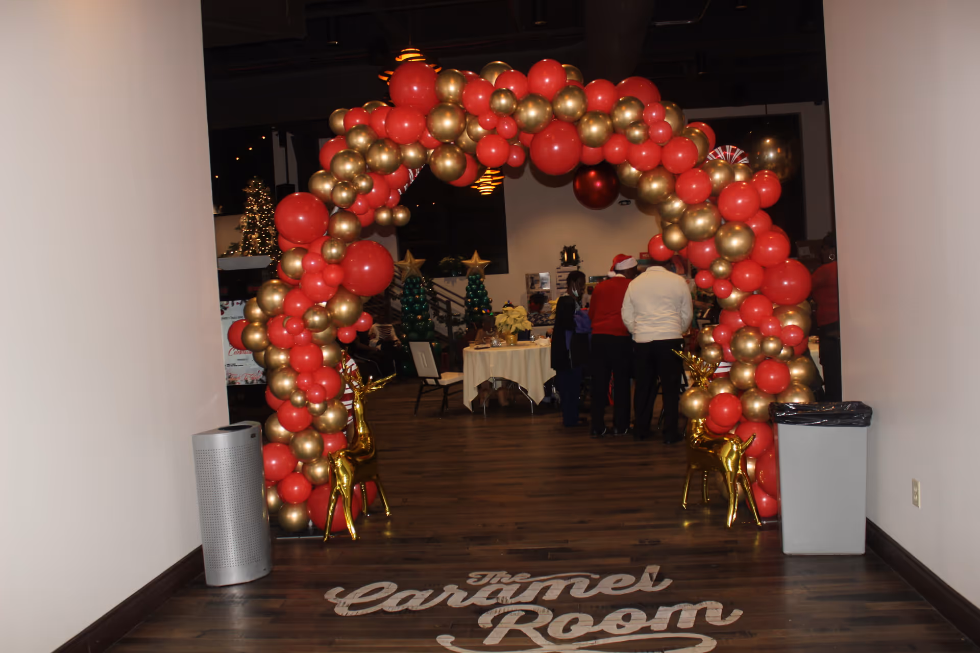 Indoor hallway with a red and gold balloon arch and holiday decorations around a table.