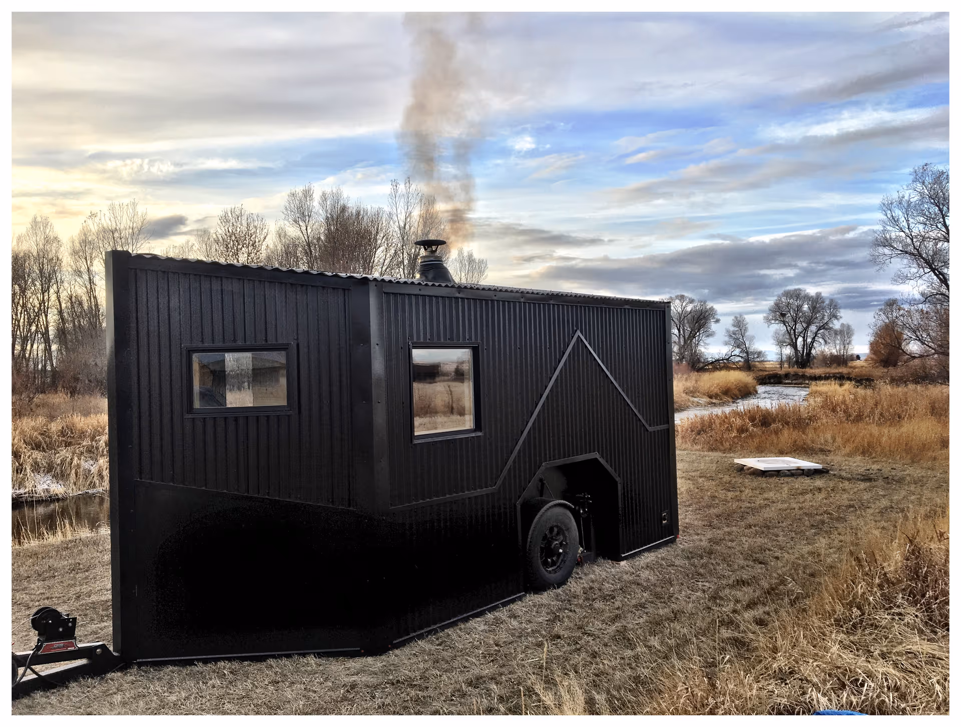 A black mobile sauna trailer sits on a grassy field beneath a cloudy sky.