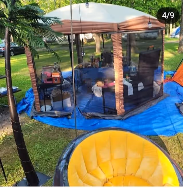 Outdoor camping tent setup with mesh walls, blue ground tarp, and chairs in a park.