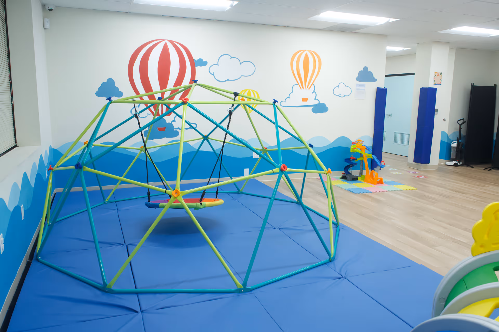 Indoor children's play area with a colorful geodesic climbing dome and padded blue mats.