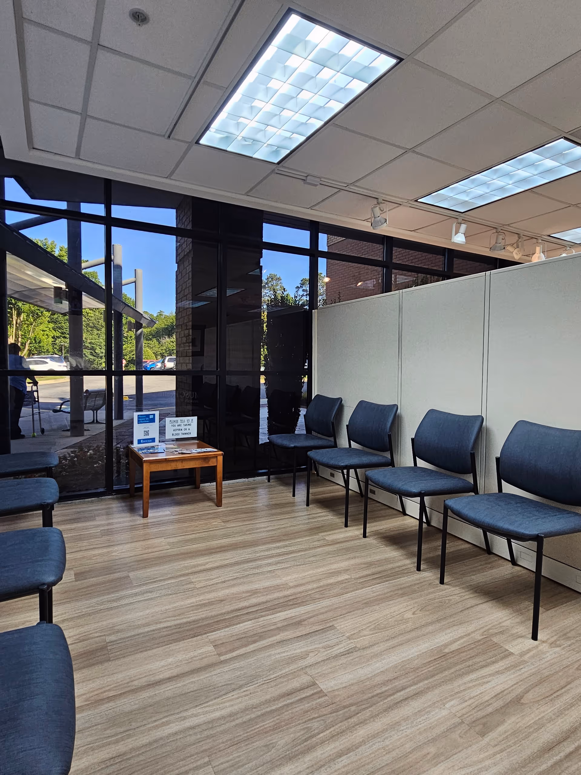 Interior waiting area with blue chairs, large glass windows, overhead lighting, and a small brochure table.