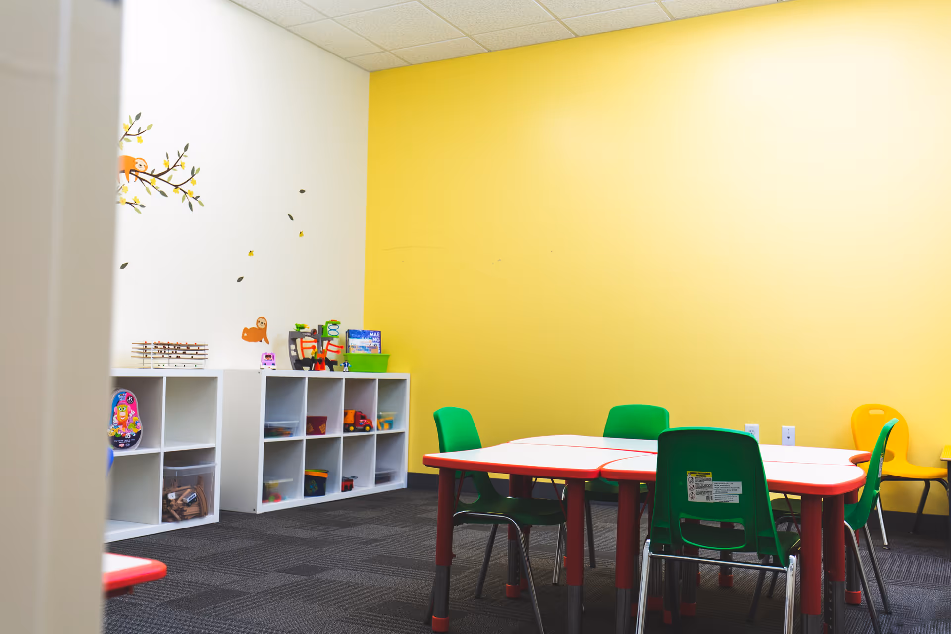 Bright yellow-walled activity room with cubby shelves, tables, and colorful chairs.