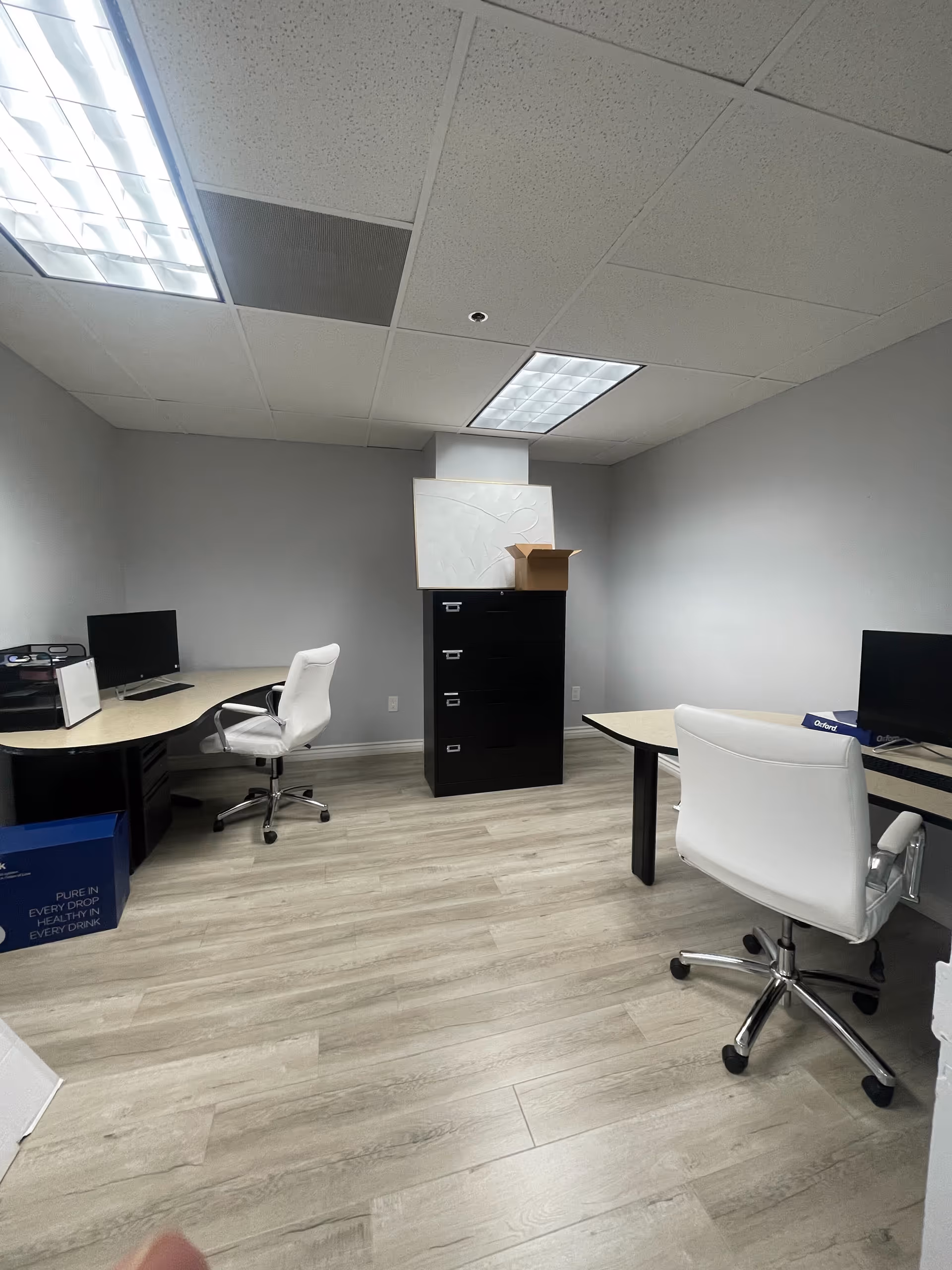An empty modern office area with desks, chairs, and a filing cabinet in a gray room.