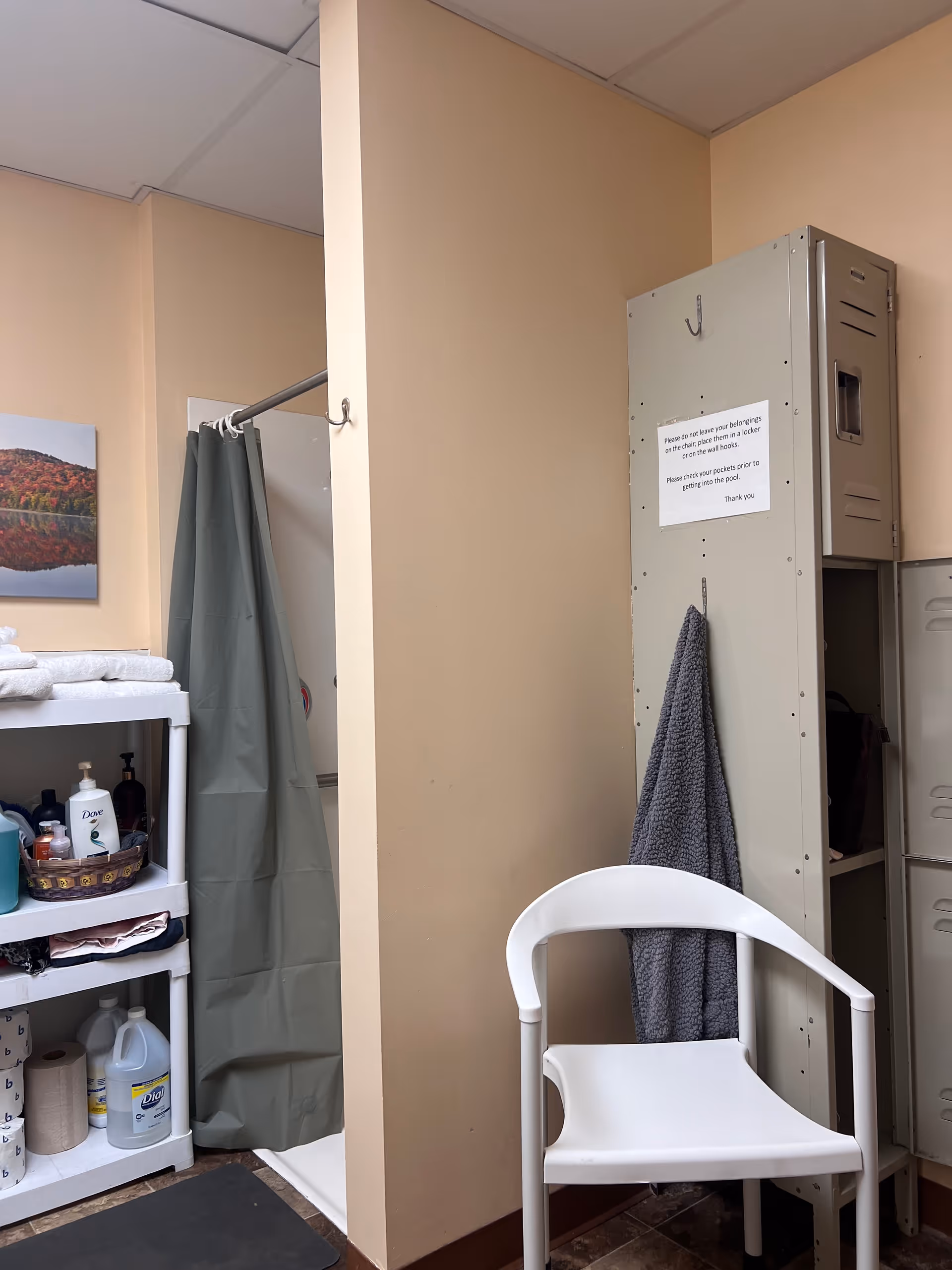 Small bathroom area with shower curtain, shelves of toiletries, a metal locker, and a white chair.