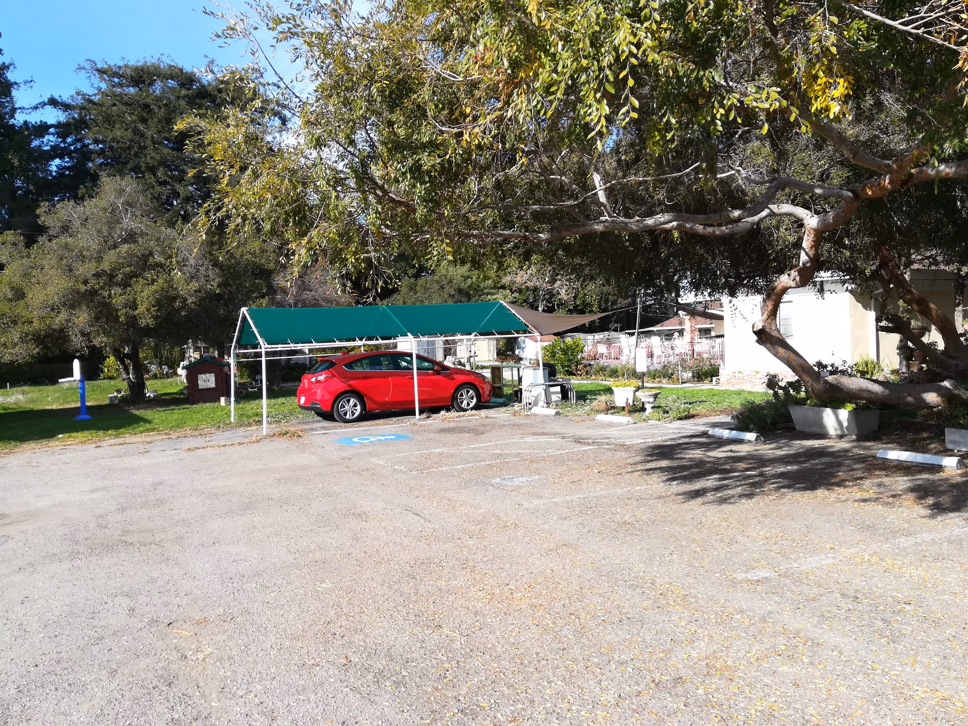 Red car parked under a green canopy beside a building with trees and parking.