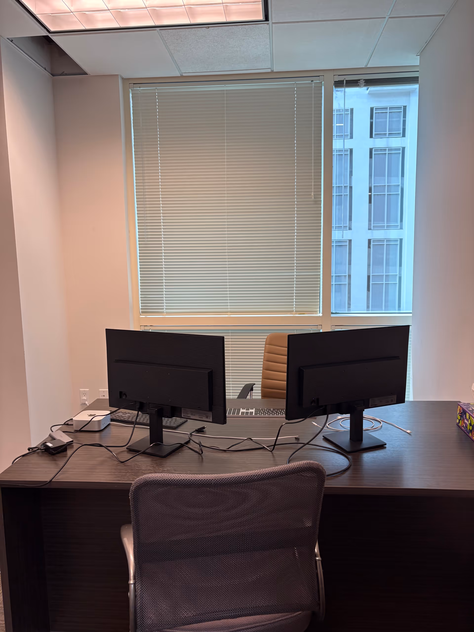 Two computer monitors on a dark desk in a windowed office with blinds.