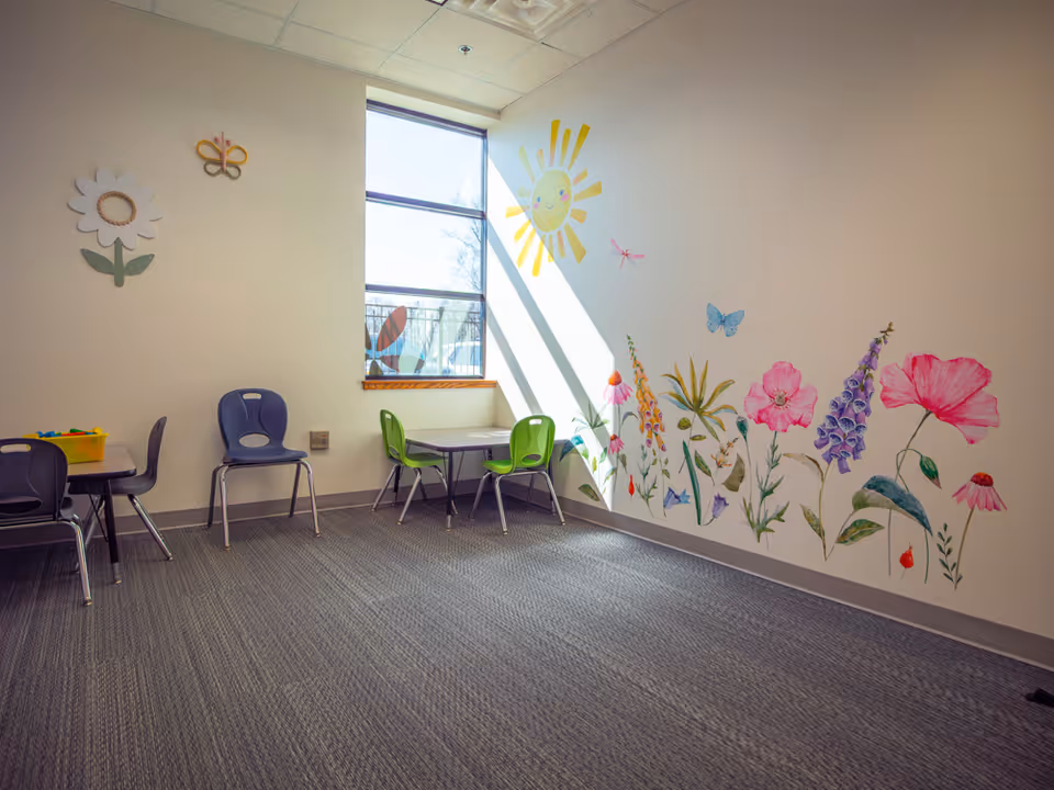 A bright activity room with a floral mural, windows, and small tables with colorful chairs.