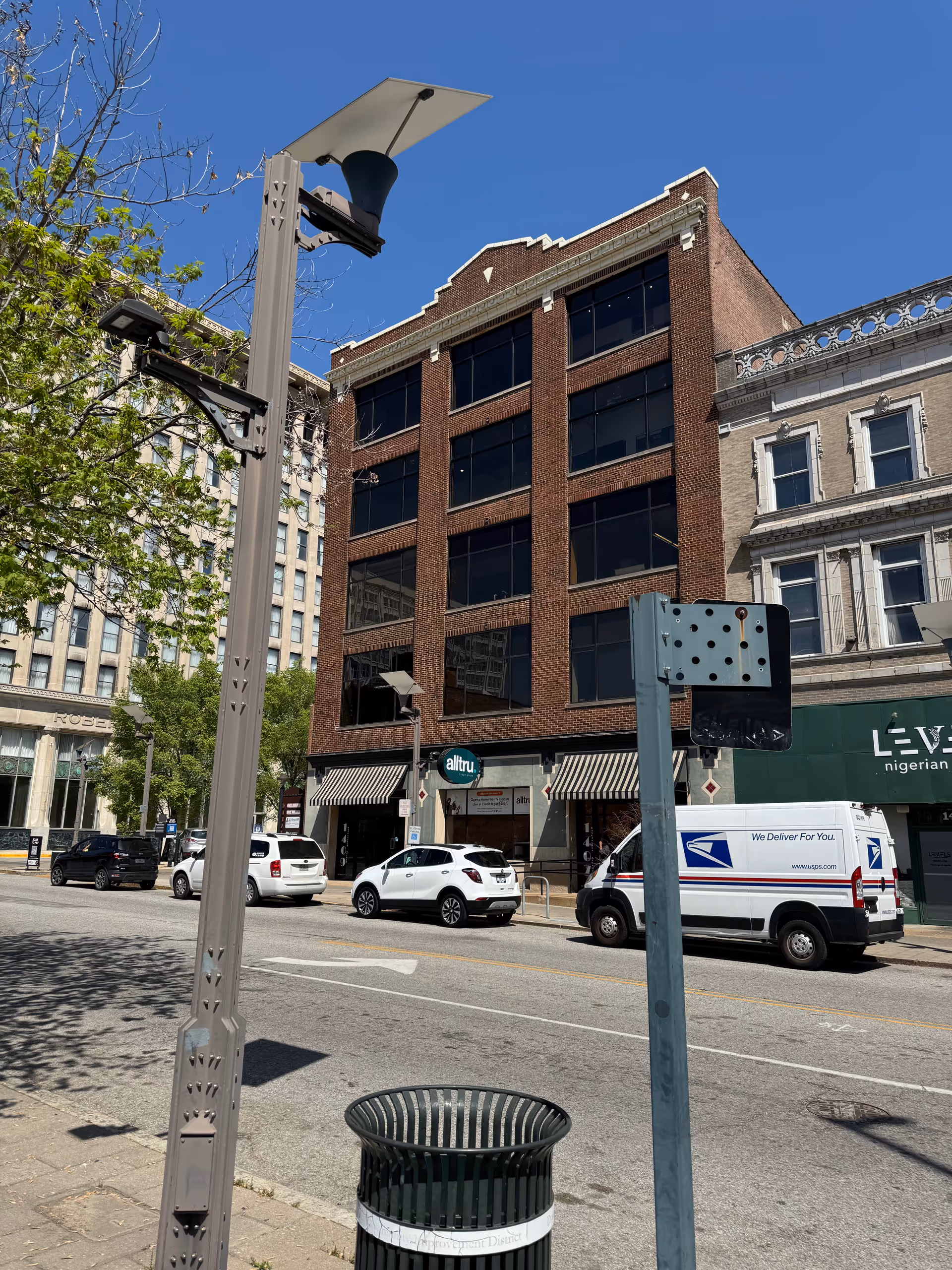 Brick multi-story building on a city street with parked cars and a trash can.