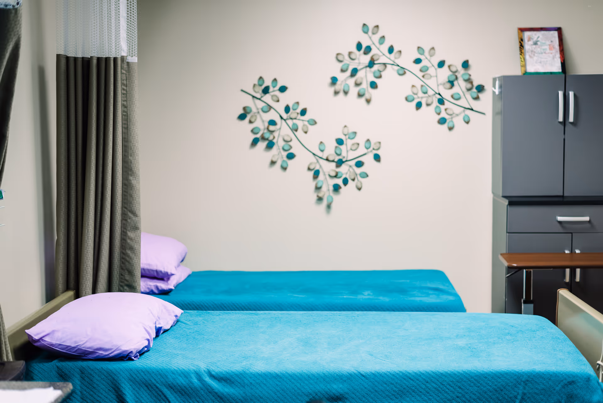 Two teal bed covers with purple pillows in a twin-bedroom, wall art, and a cabinet.