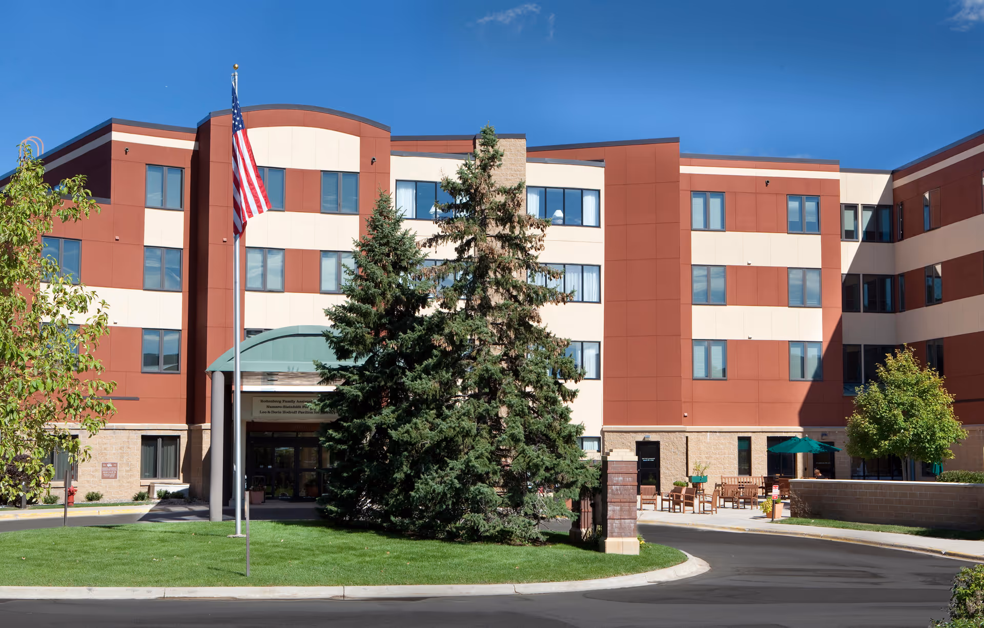 Exterior view of a multi-story senior living building with flag, trees, and outdoor seating.