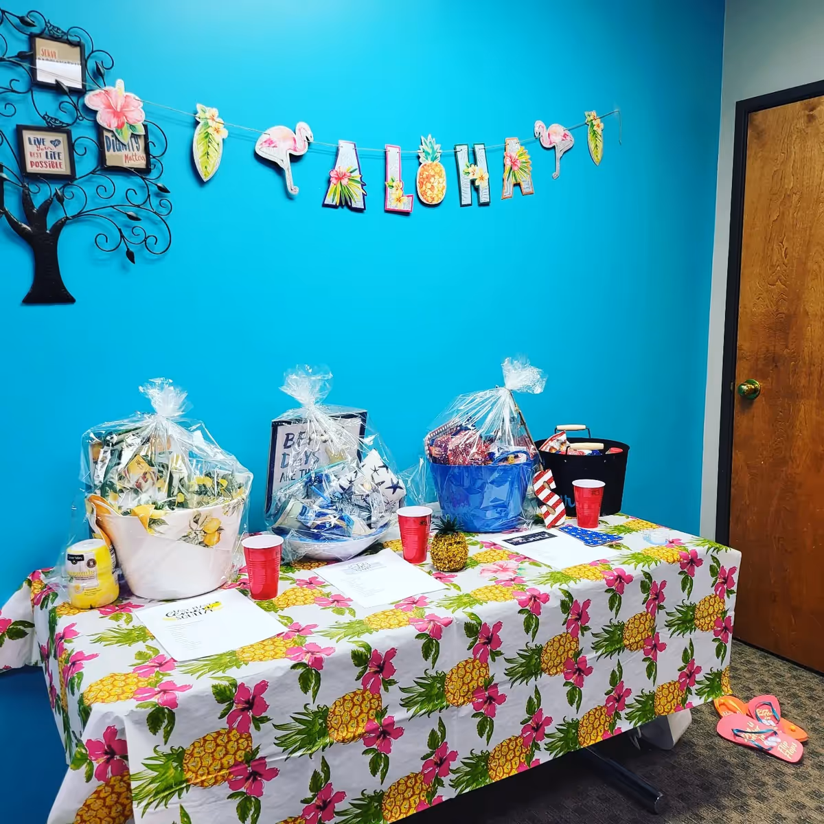 Bright turquoise wall with tropical-themed table, pineapple decor, gift baskets, and an Aloha banner indoors.