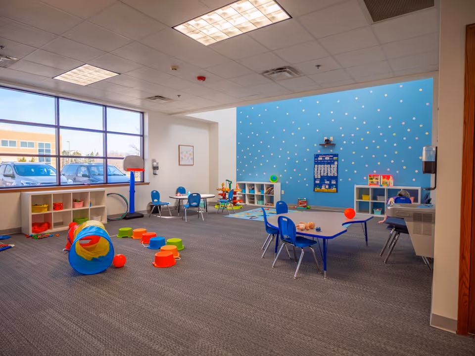 Bright indoor playroom with blue wall, toy shelves, small tables, chairs, and play equipment.