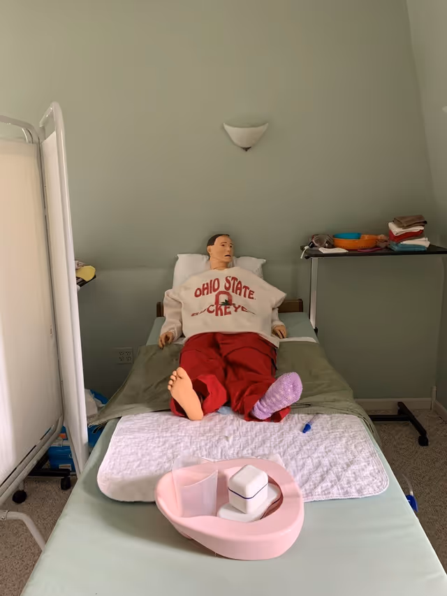 A medical training mannequin lies on a bed in a small care room with supplies.