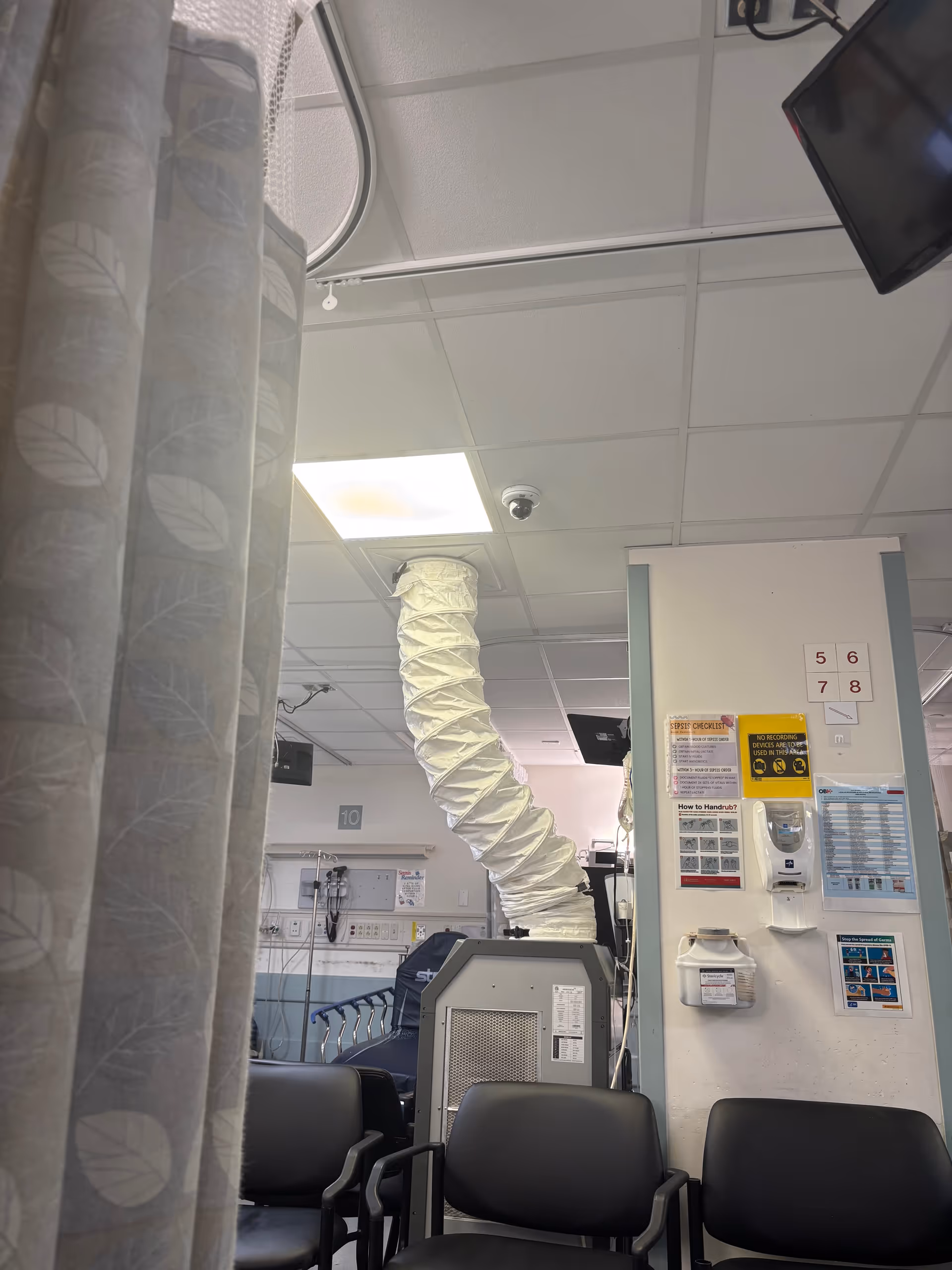 Hospital interior with privacy curtains, seating, and a large flexible white ventilation duct.