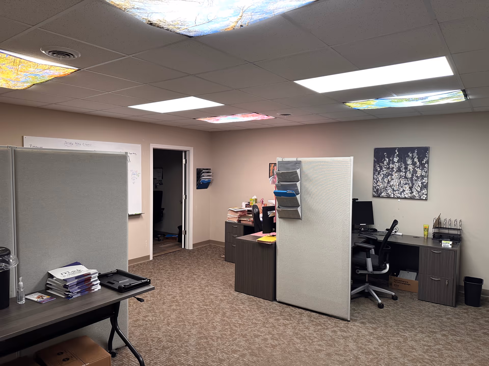 Office area with cubicles, desks, and paperwork in a neutral room inside a care facility.