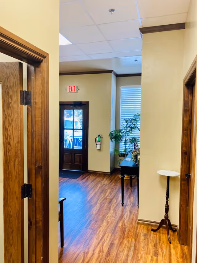 Interior hallway of a senior living facility with doors, exit sign, plants, and wooden floors.