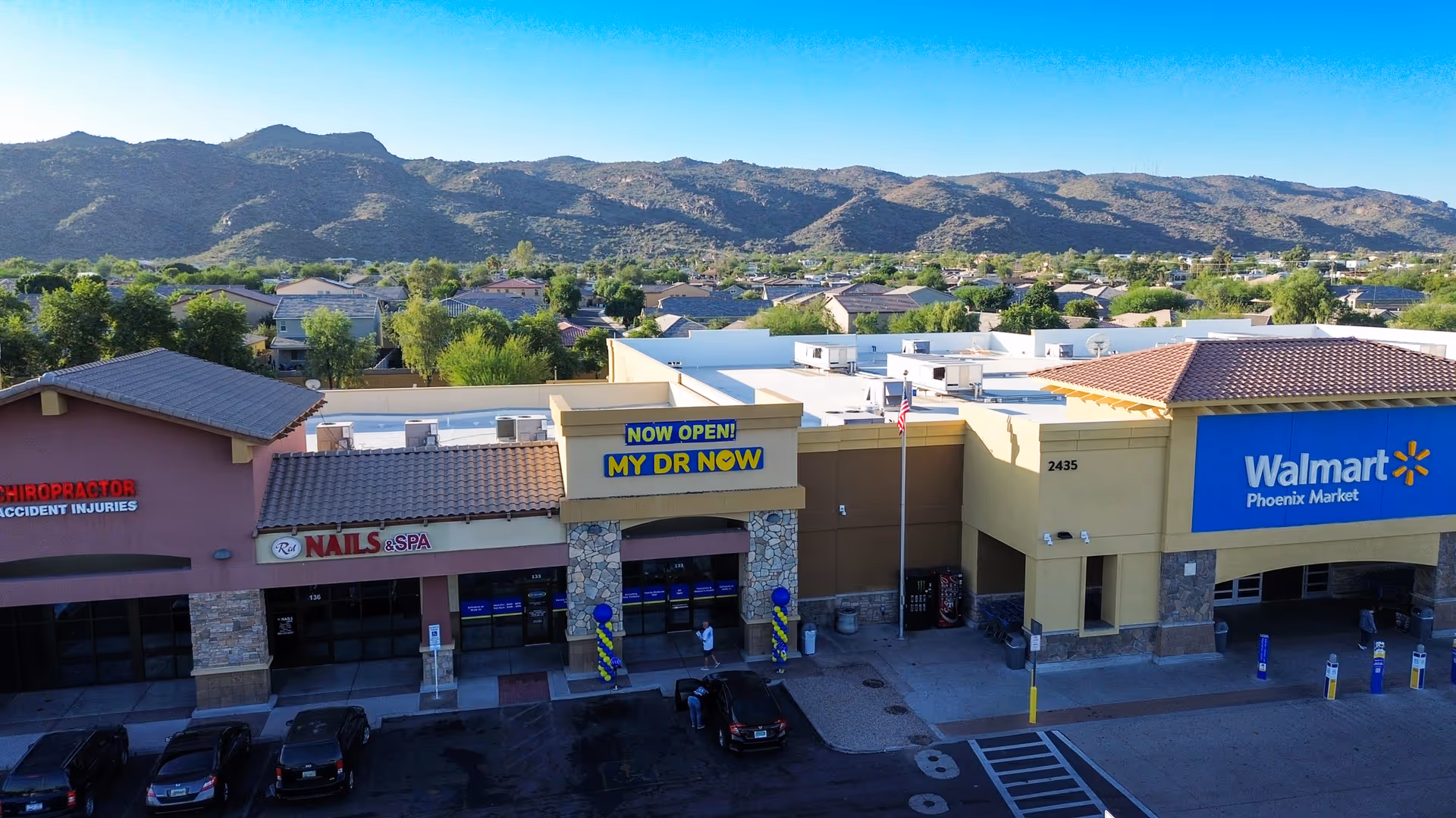 Exterior of a strip mall with MY DR NOW between Nails Spa and Walmart, mountains behind.