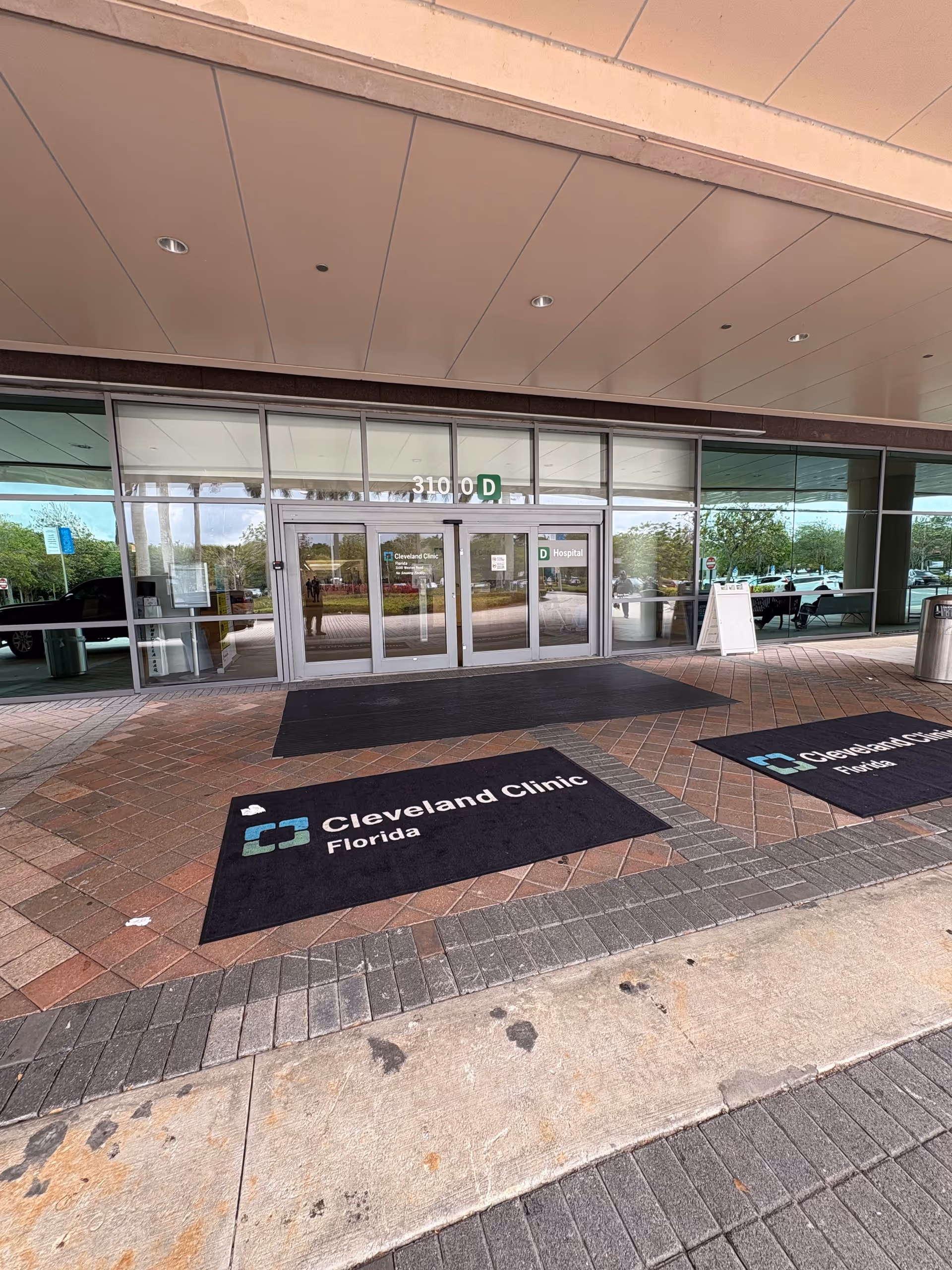 Glass-front entrance with Cleveland Clinic Florida mats and a brick-paved walkway.
