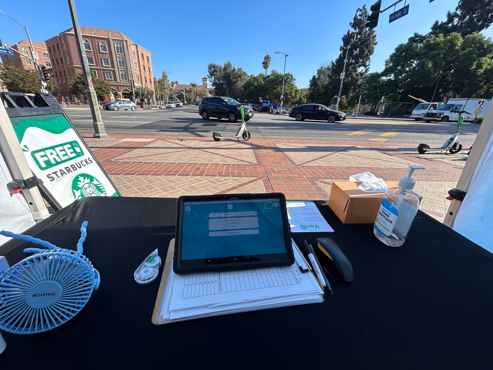 Outdoor tent setup with tablet, paperwork, sanitizer, hand gel, and supplies on a black table.