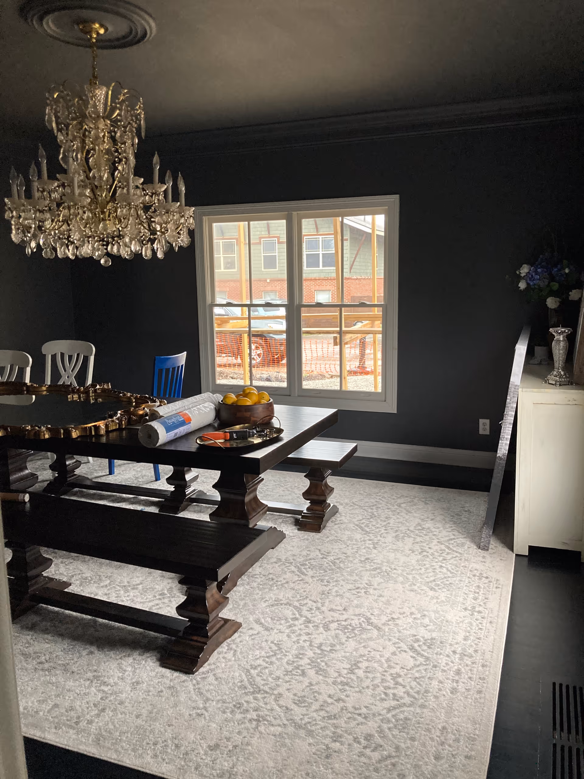Dark-walled dining room with crystal chandelier, large wooden table, benches, and a window view.